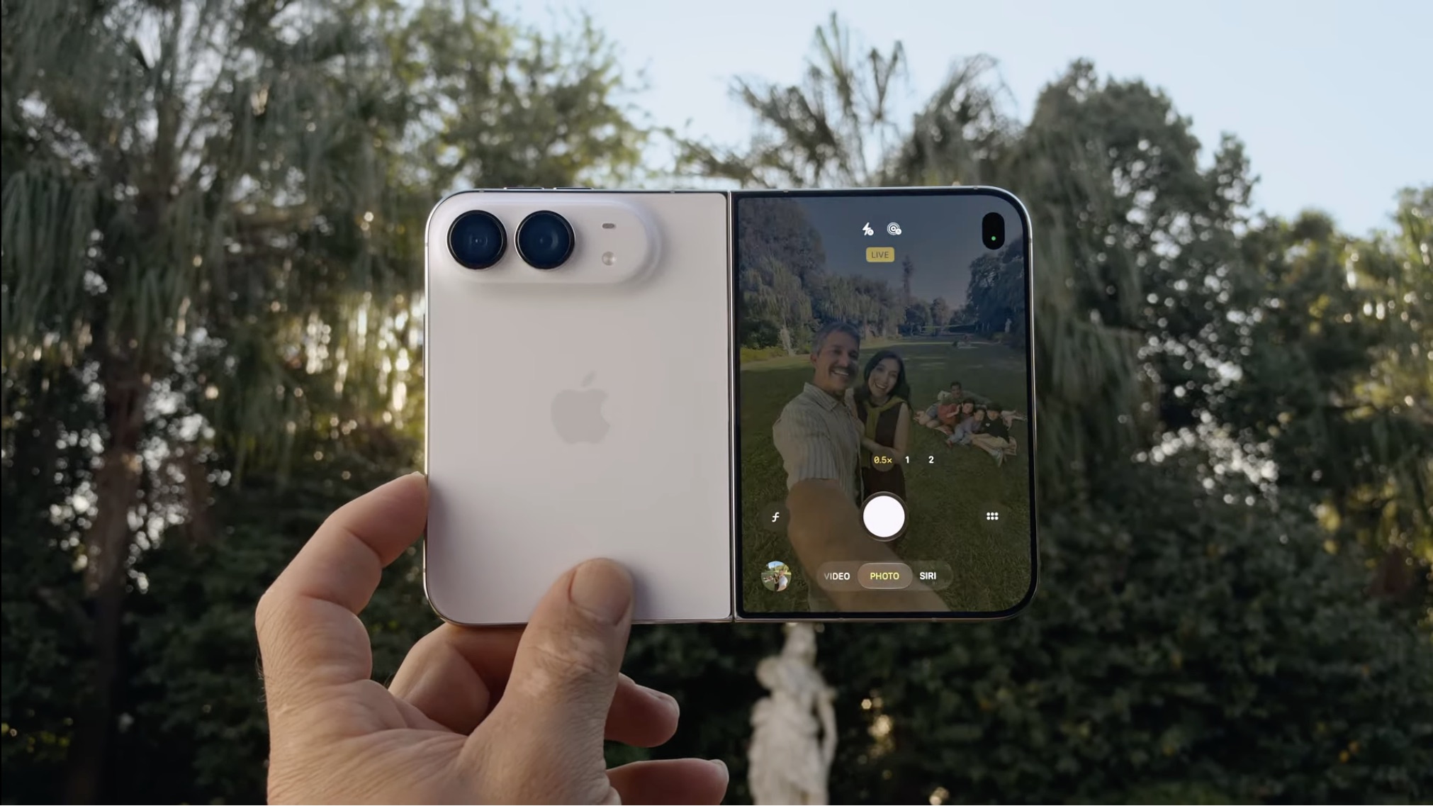 Uma mão segura um smartphone dobrável branco, mostrando a tela externa com o aplicativo de câmera aberto, exibindo uma prévia de selfie com pessoas ao fundo.