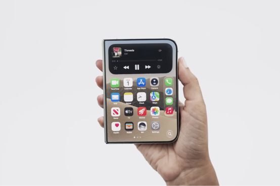 Uma mão segura um smartphone dobrável fechado, exibindo a tela externa com ícones de aplicativos e um widget de mídia no topo, contra um fundo branco neutro.