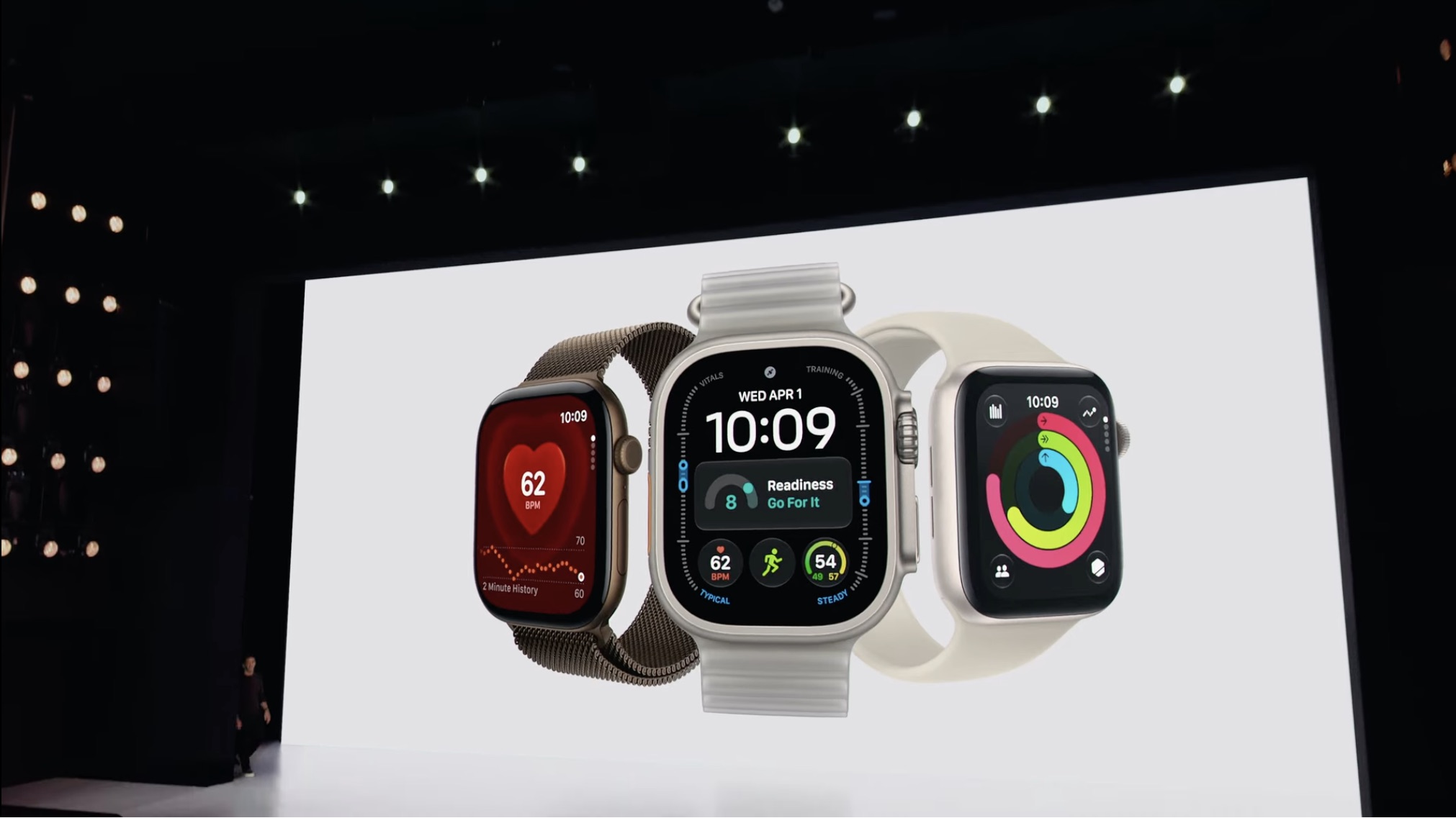 Três modelos de Apple Watch exibidos em uma tela grande, destacando mostradores com dados de frequência cardíaca e monitoramento de saúde e atividade física.