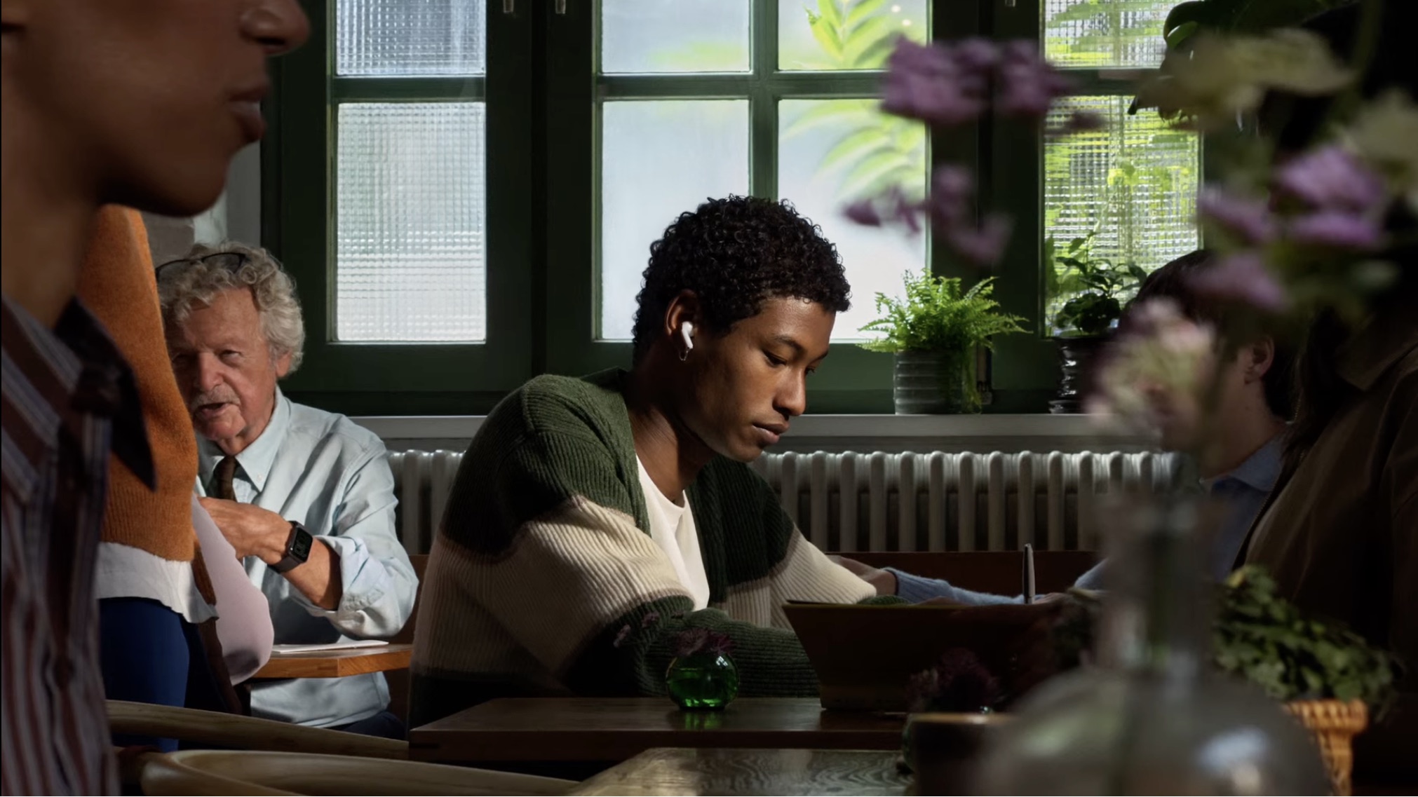 Um jovem usando AirPods nas orelhas está sentado em um ambiente interno com outras pessoas ao redor.