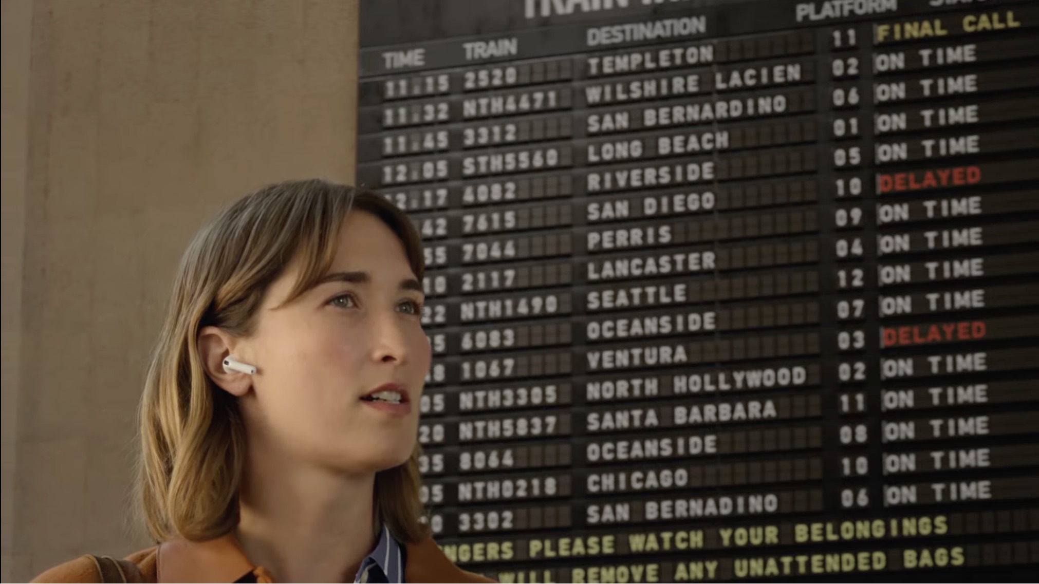 Uma mulher usando fones AirPods em frente ao painel de horários de trens em uma estação.