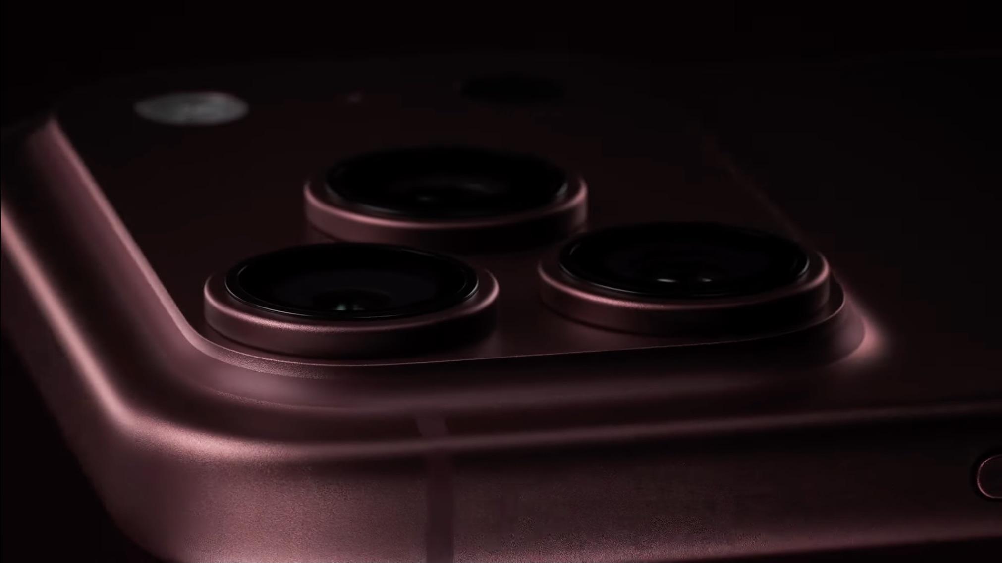 Close-up do módulo de três câmeras traseiras de um smartphone na cor borgonha, com iluminação suave em fundo escuro.